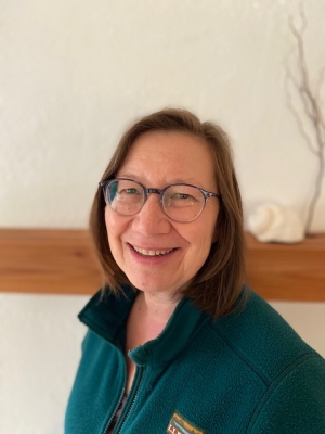 headshot photo of middle-aged Asian-American woman wearing purple rimmed glasses and a teal LL Bean fleece. She is smiling and looking at the camera. She has shoulder length brown hair.