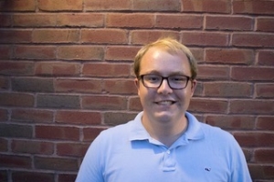 Photo of young male with blonde hair wearing brown-rimmed glasses, smiling in a light blue photo shirt against a brick background.