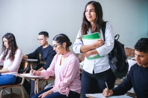 A classroom of adult students, one standing with a book in her arms, as the other students look down at their desks