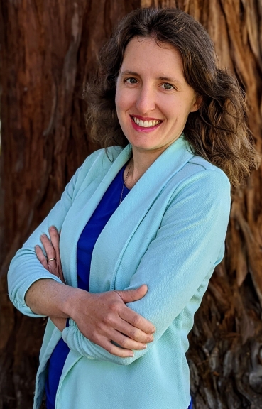 Professor Yelena Proskurin stands smiling with arms crossed in a light blue blazer in front of a tree.
