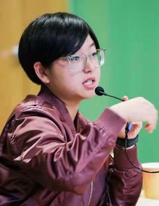 Professor Shen, with short dark hair, glasses, and a red jacket, stands at a microphone mid-speech, gesturing with her hands and looking toward an off-camera audience.