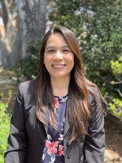 Professor Matsuda stands smiling into the camera with long brown hair, a black blazer, and floral shirt with trees in the background.