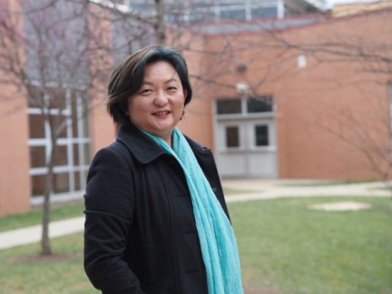 Professor Eu stands in front of a building in a black blazer, blue scarf, and short dark hair.