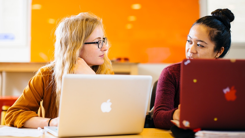 Two students work side-by-side on computers in the brightly lit student lounge.