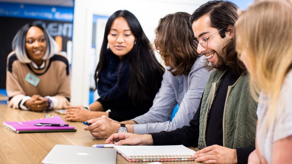 Five students sitting together at a long table have a discussion.