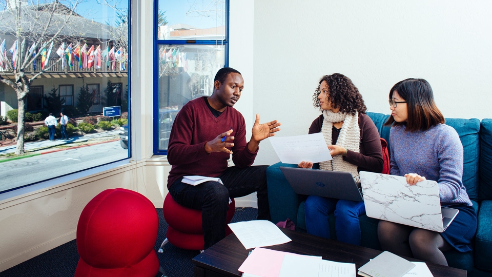 Four students sit at a table in front of a picture window having a discussion.