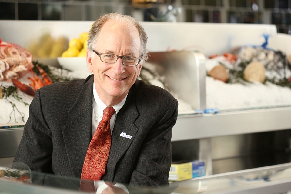Mr. Andy Sharpless, smiling, in a business suit leaning on a counter, behind him is a fish market counter with various fish and seafood on offer