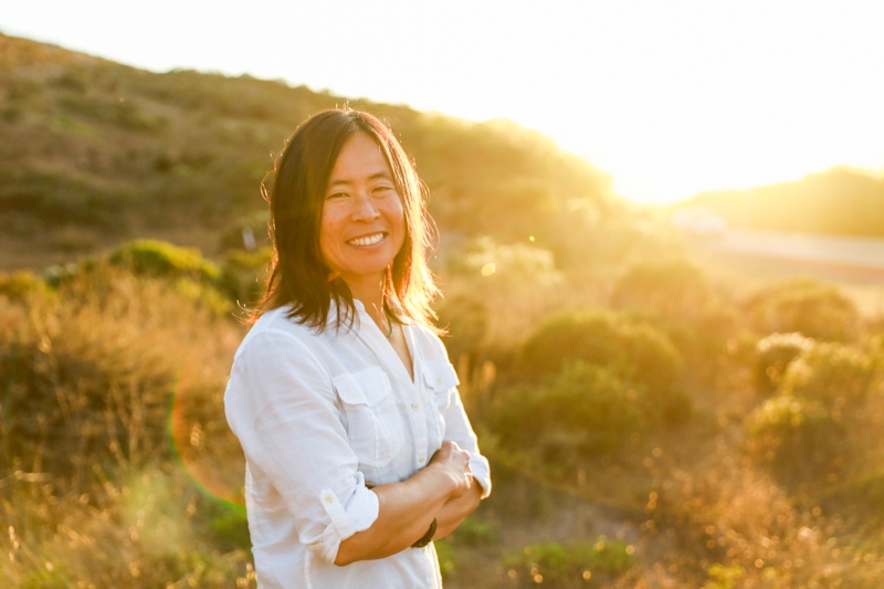 A smiling Miho Aida with arms crossed in beautiful outdoor setting grassy hills with sunlight in background