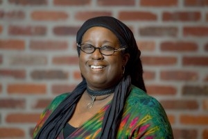 Jacqueline Patterson headshot, she is smiling and wearing glasses, dressed stylishly and posing against a brick wall