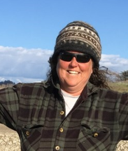 Monique Fountain, Director of the Tidal Wetland Program at the Elkhorn Slough National Estuarine Research Reserve.  She is wearing a warm, knit cap and flannel shirt, smiling brightly with red cheeks and sunglasses against a blue sky with clouds, and the greens and browns of the slough.