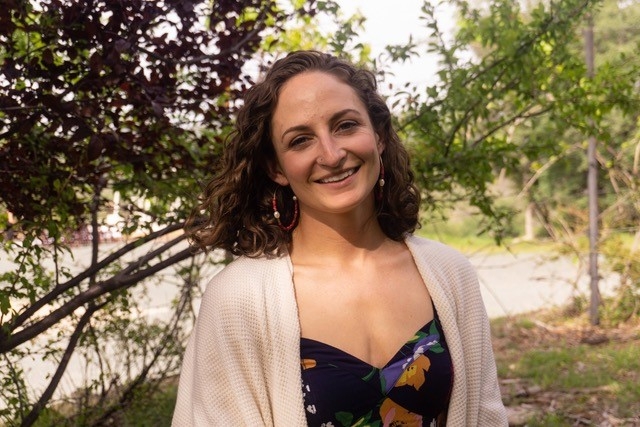 Ella McDougall, smiling with kind eyes, shoulder length curly brown hair, standing against a backdrop of trees