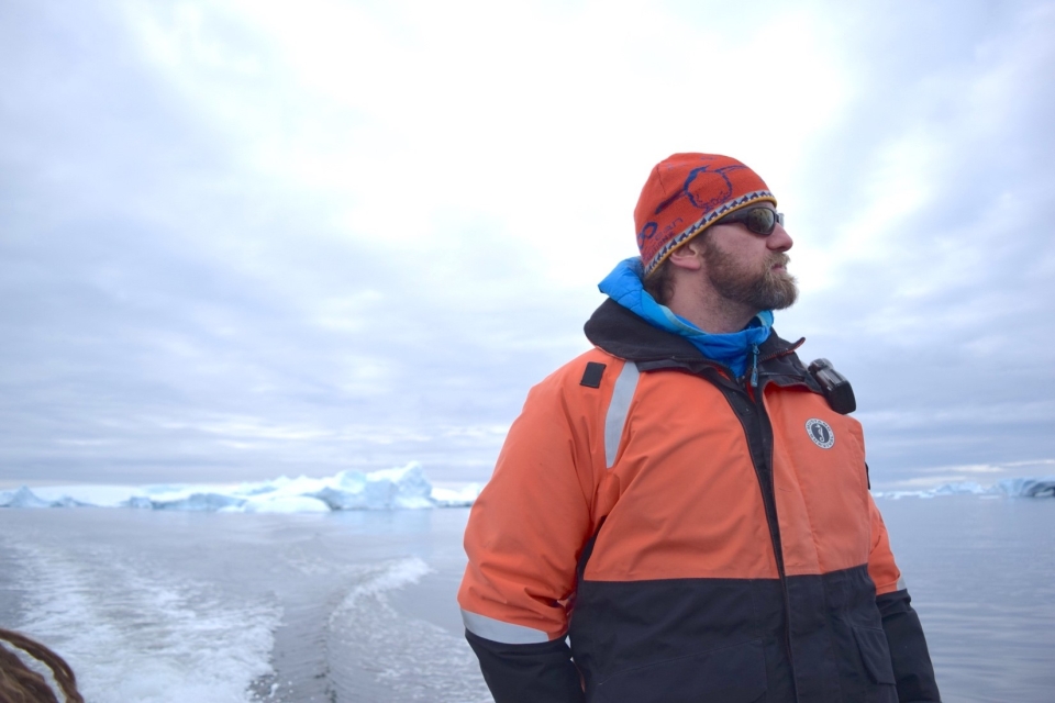 Ari S. Friedlaender, PhD, looking badazz in profile against an Arctic sky--he studies whales and is awesome. White/blue sky, white/blue glacial ice and ocean water, he is wearing an orange/black jacket, red snug cap, and sunglasses.
