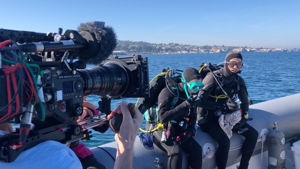 A film crew shooting divers about to go overboard with dive gear on--blue ocean and bay behind