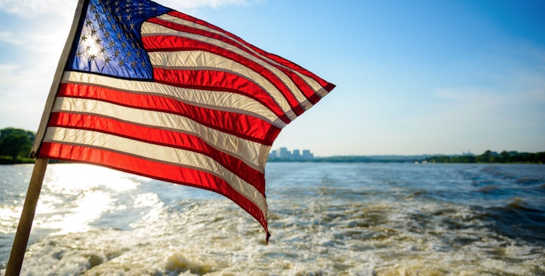 the us flag at the stern of a vessle on the ocean, overlooking water and city skyline