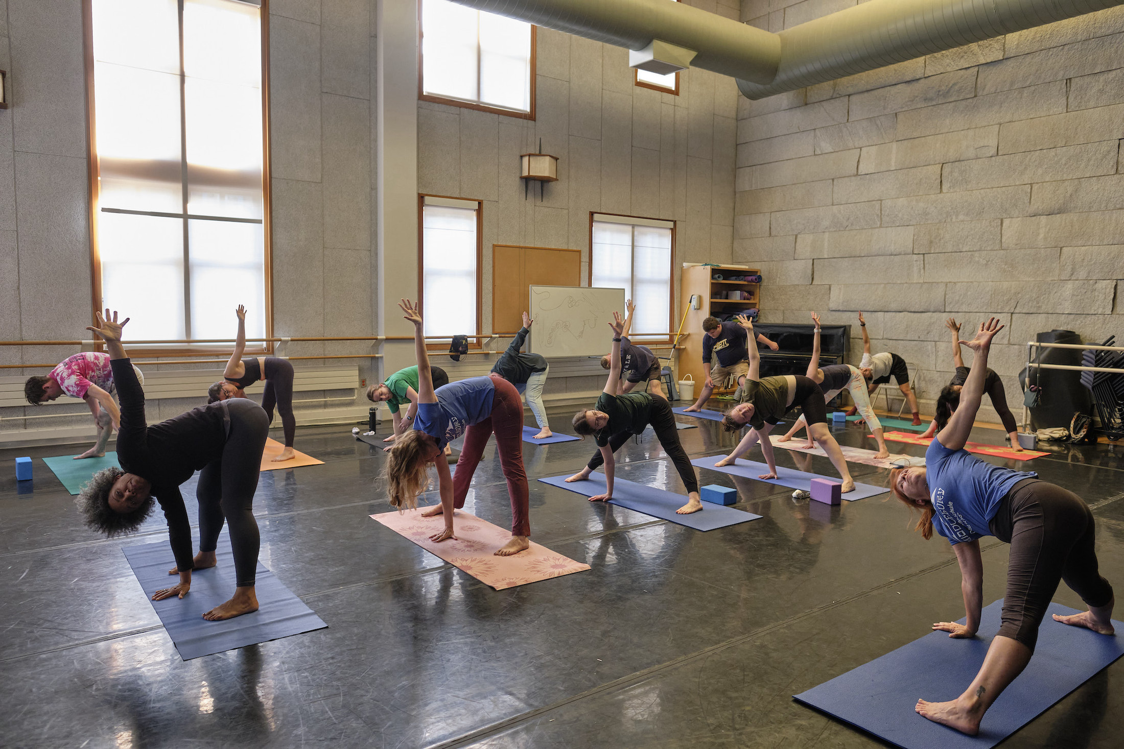 A group of people does yoga in a well lit room- star pose. 