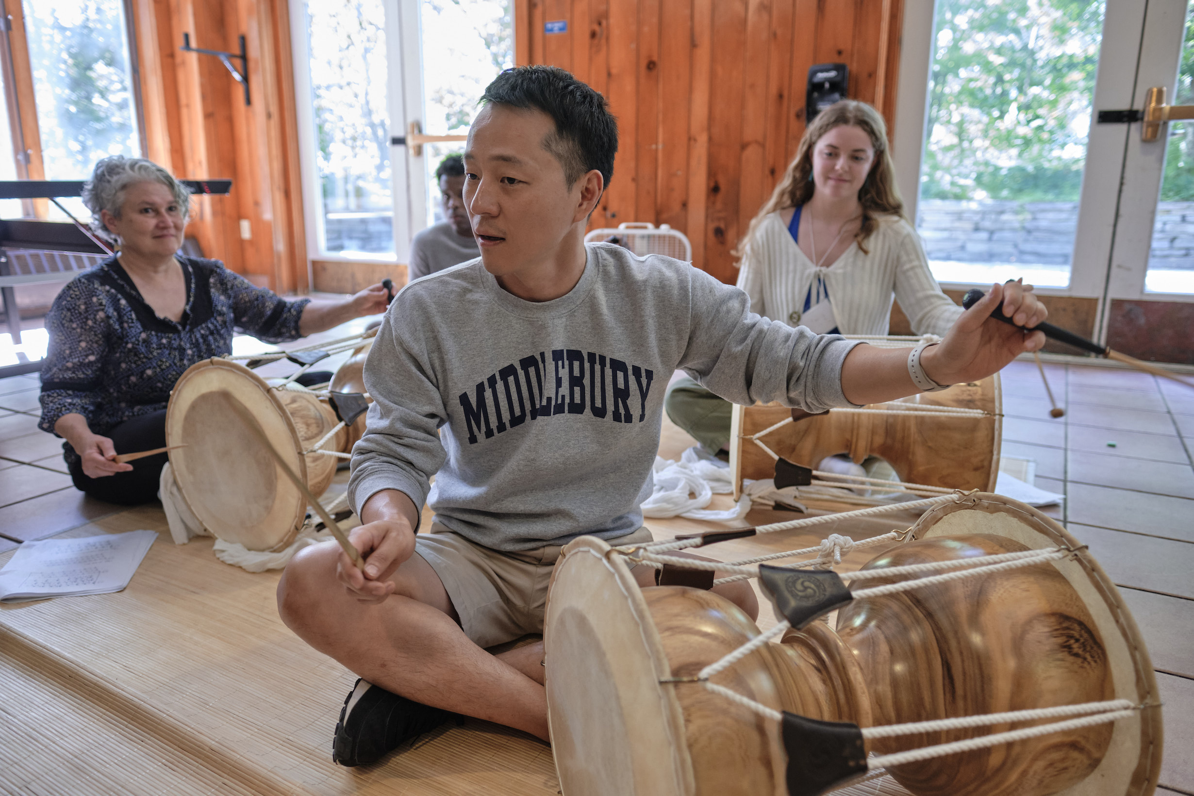 A teacher teaches students how to drum on a Korean drum. He sits in front of his students, leading them.