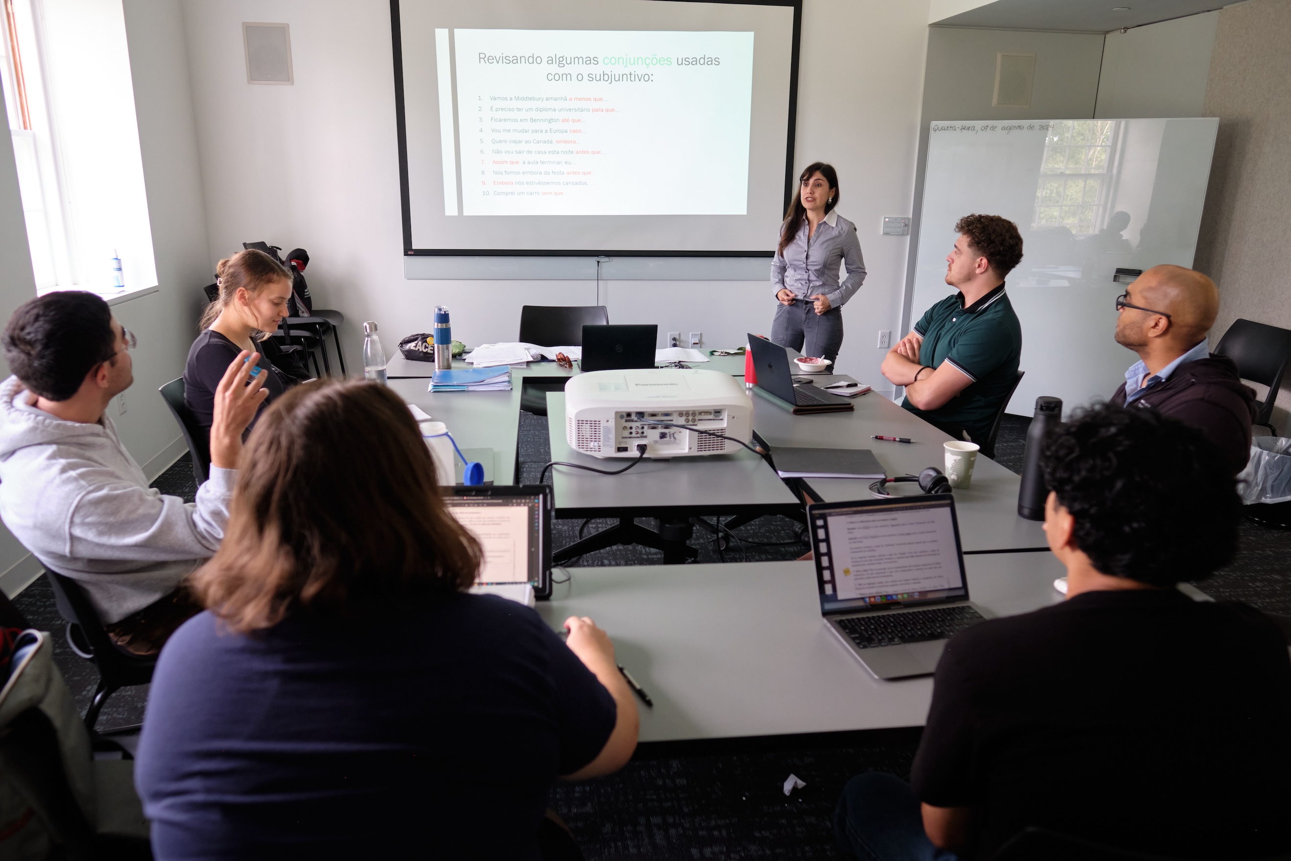 A portuguese teacher speaks with her students, standing in front of a projector screen.