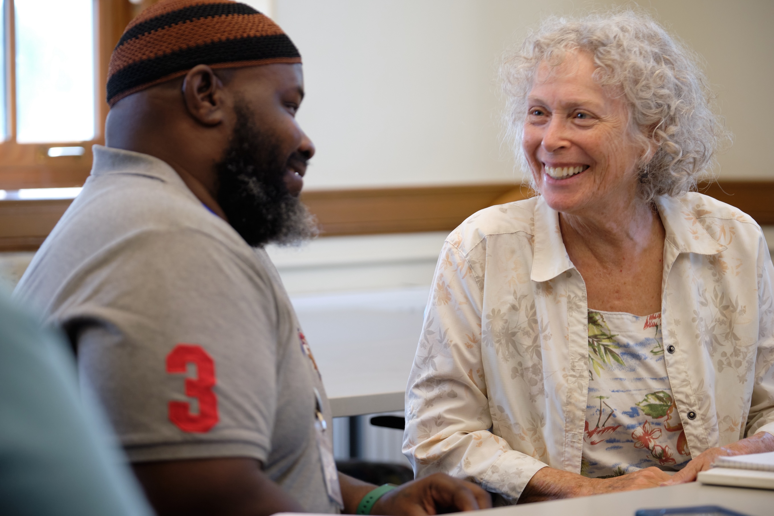 Two students sit in class, smiling as they chat. 