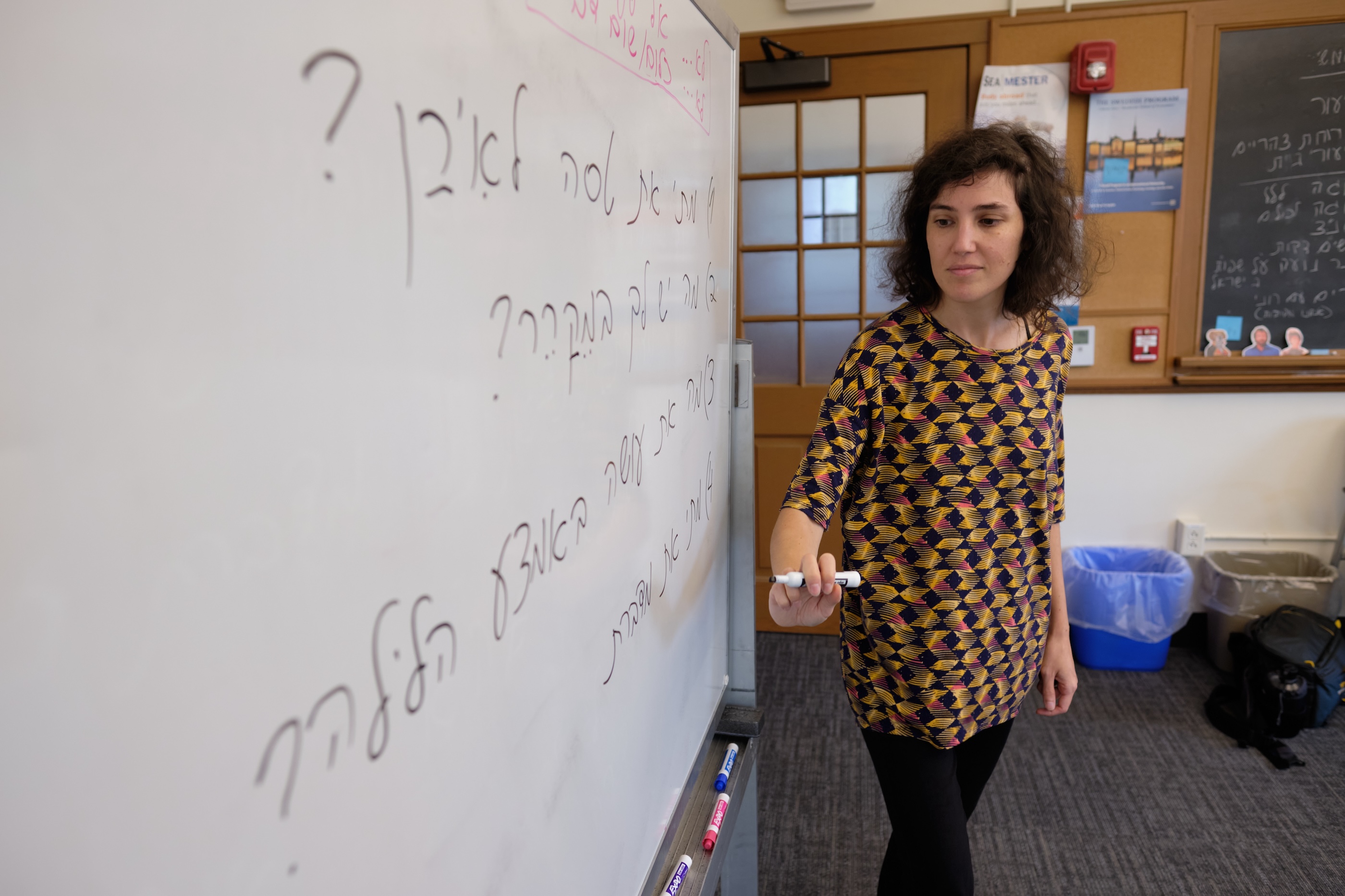 A teacher stands at the whiteboard, writing in Hebrew. 