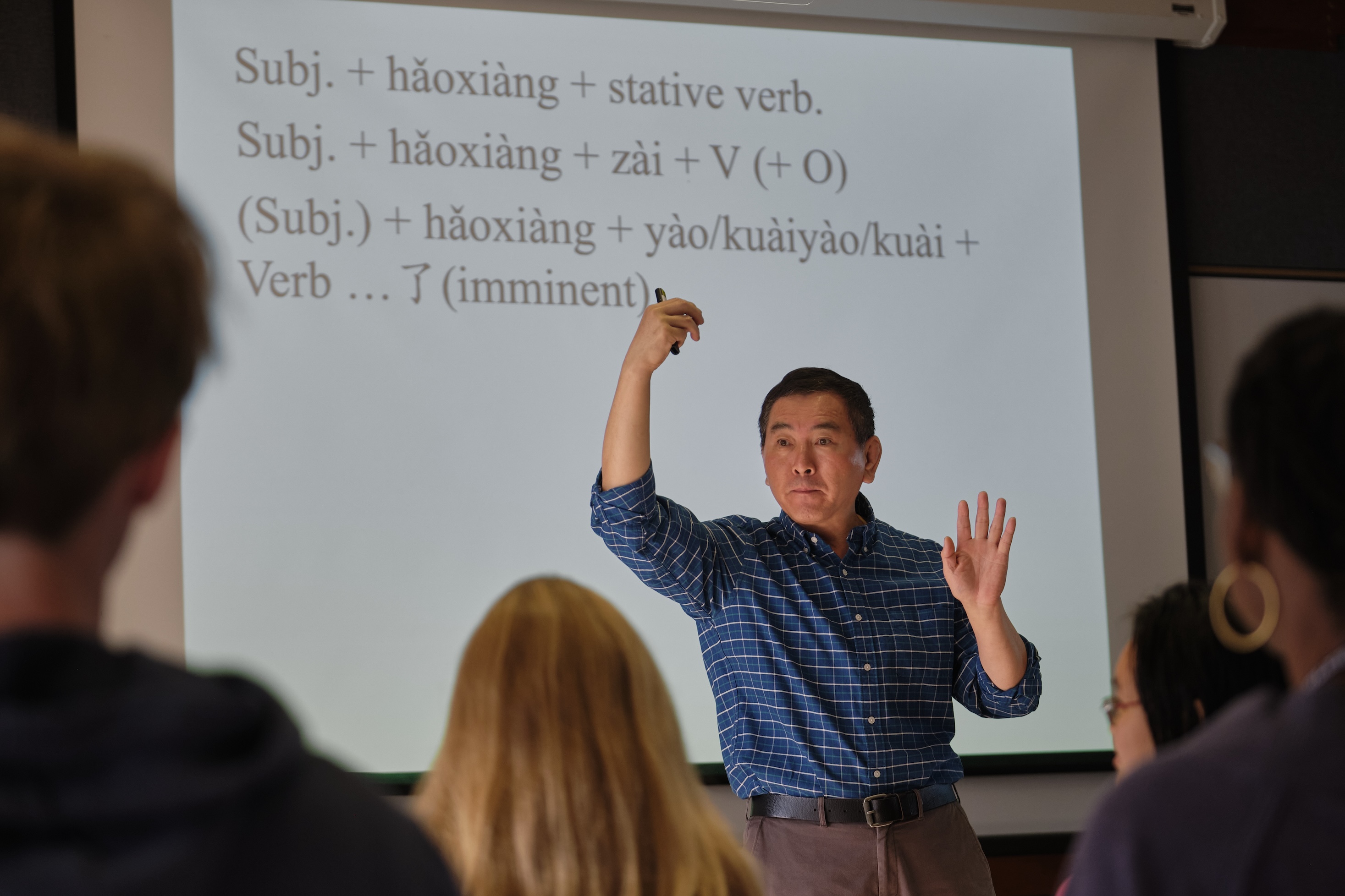 A man gestures to the Chinese grammar on a white board behind him. 