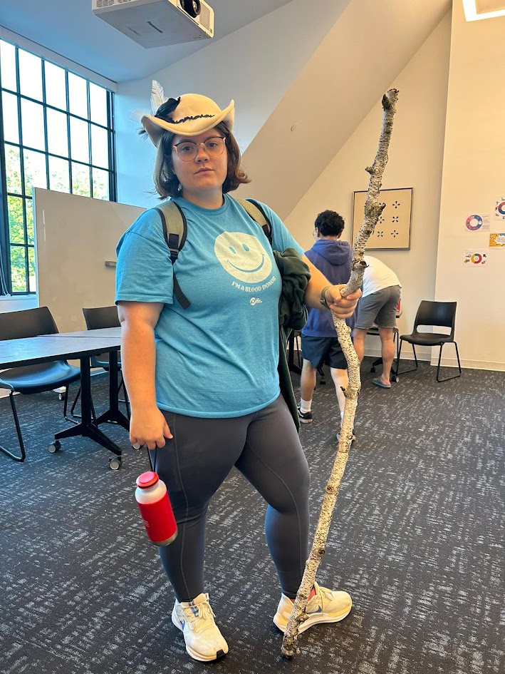 A woman in a blue t-shirt stands with a walking stick and a hat. 