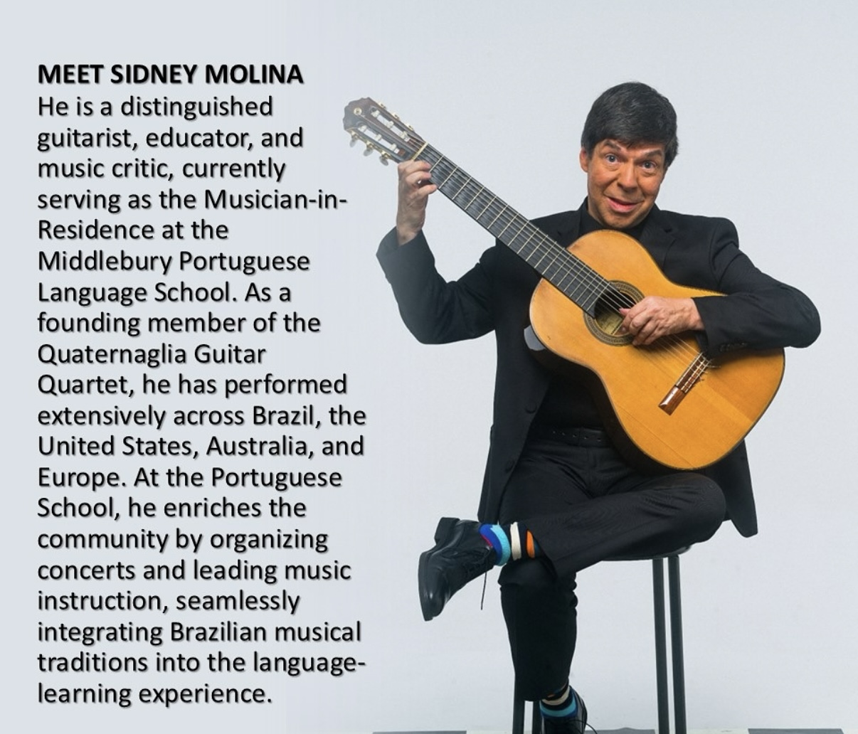 A man in a black suit with a guitar smiles at the camera. 