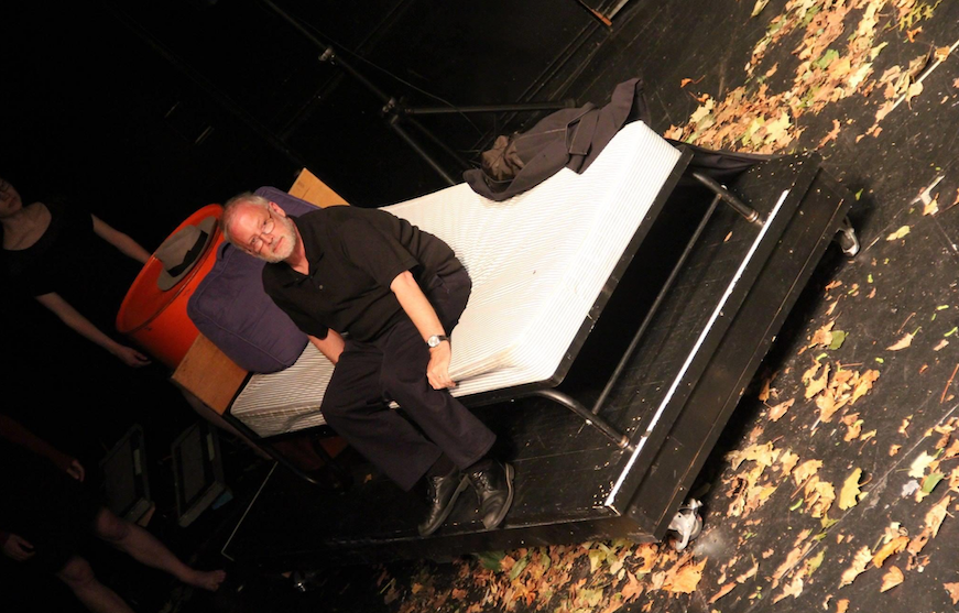 A man in a black shirt sits at the end of the bed in a School of French play at Middlebury.
