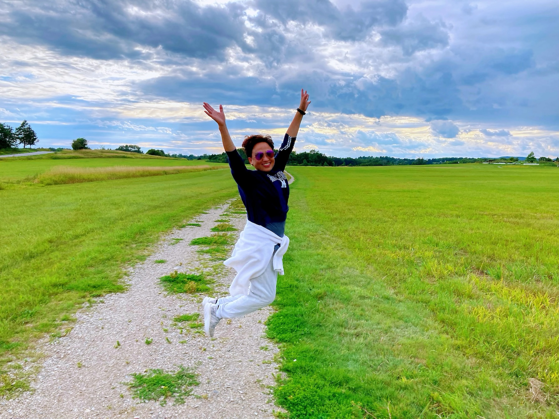 A woman in a black shirt and white jacket around her waist jumps for joy in a field