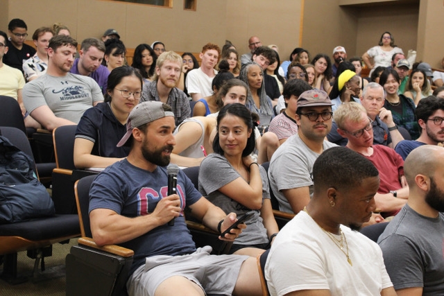 Students sit in the audience in an auditorium, one student speaks into a microphone