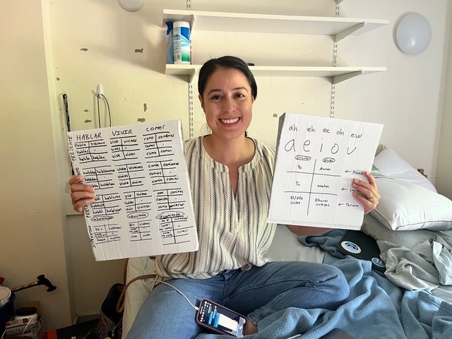 A woman holds up white boards in her dorm room with Spanish grammar practice written on them