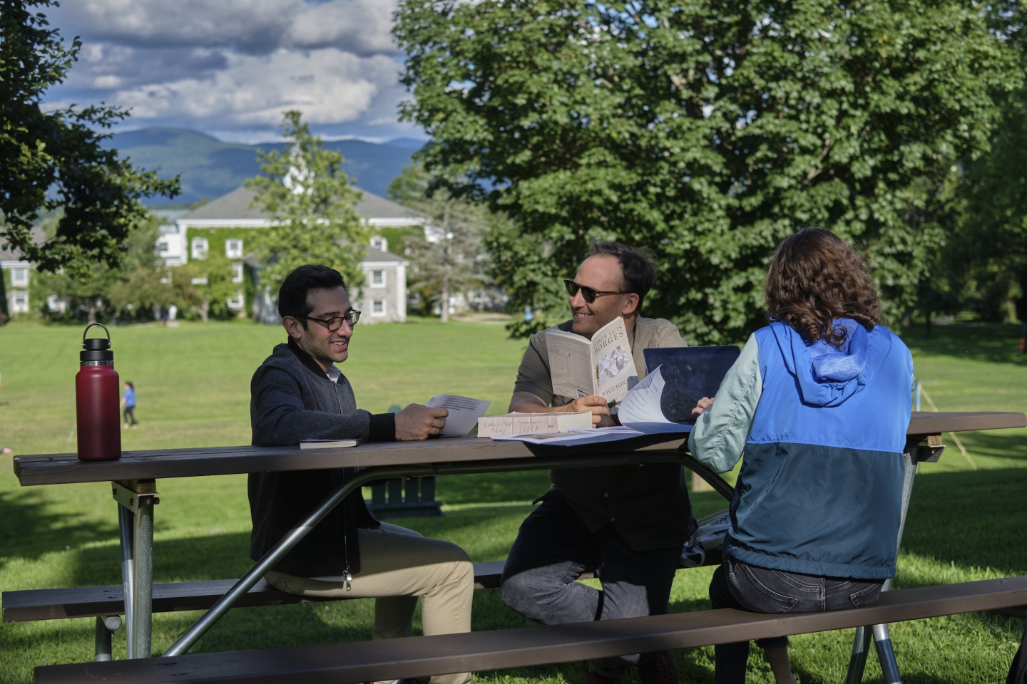 Students smile as they discuss a book, sitting outside at a picnic table. 