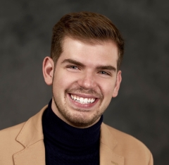 A man in a black turtleneck and tan jacket smiles at the camera