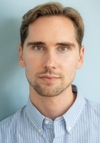 A man in a light blue button up looks at the camera. 