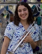 A woman in a blue and white blouse holding a trumpet smiles at the camera.