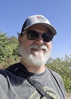 A man in a baseball cap and sunglasses smiles at the camera. 