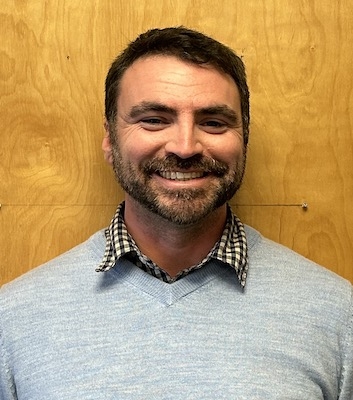 A man smiles at the camera against a wooden wall. 