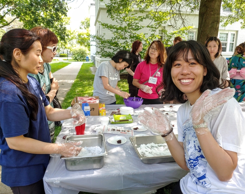 A student lifts her gloved hands into the air and smiles at the camera. She sits at a table making food.