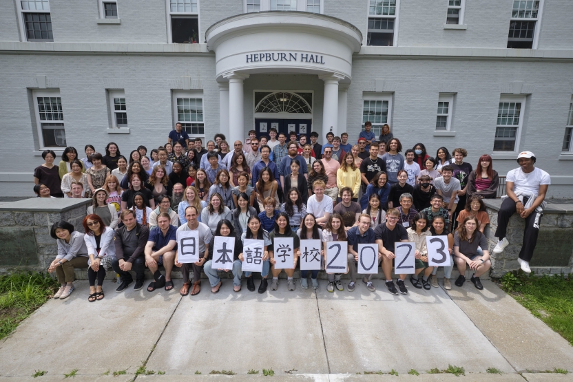 A group of people sit on the steps in front of a dorm building, holding up signs spelling out 2023 in Japanese.