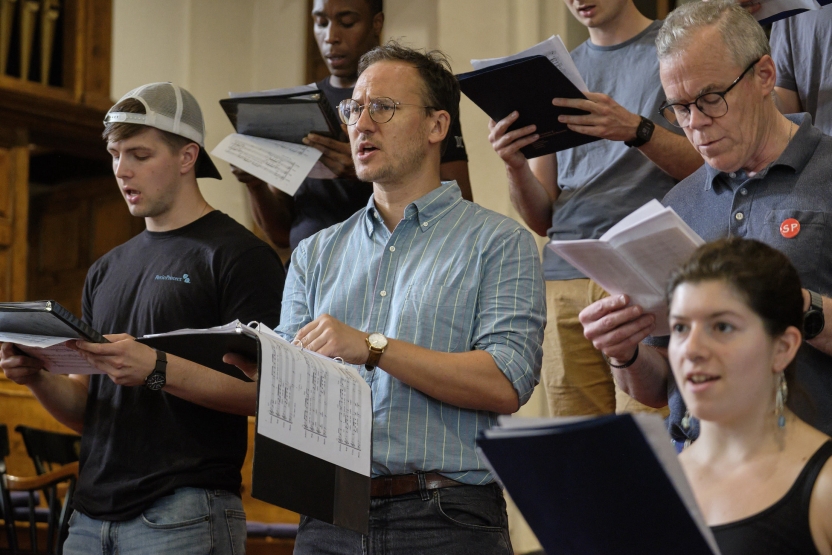 Three men and a woman stand in rows, singing in a choir in a chapel.