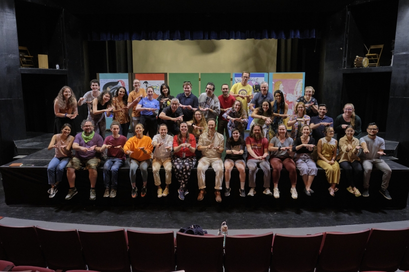 A large group of people sits on a stage smiling at the camera and making the same hand gesture.