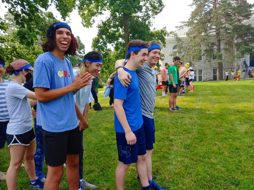 A student smiles at the camera, clapping. His team mates walk away with their arms around each other.