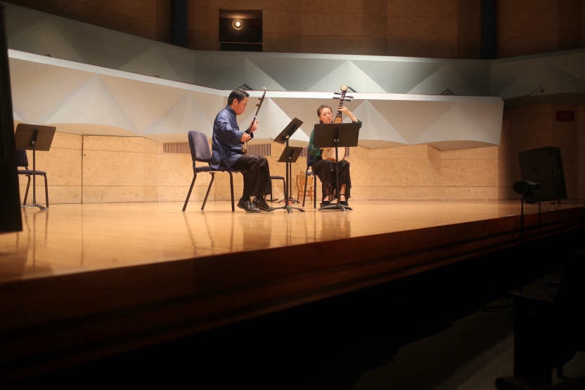 Musicians sit playing Chinese instruments on a well lit stage. 