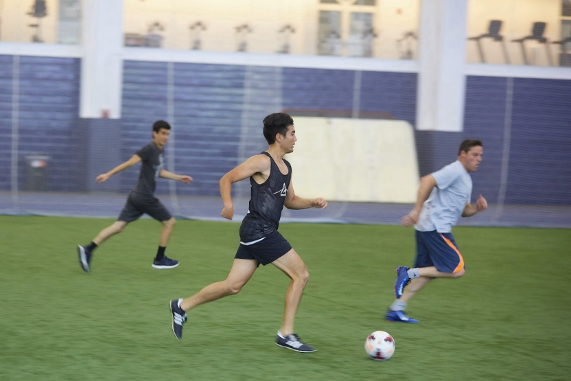 Students run across an indoor soccer field, chasing the ball 