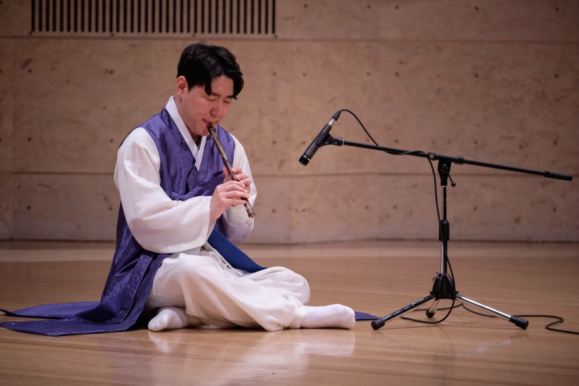 A musician plays a traditional Korean wind instrument, sitting on a stage. 