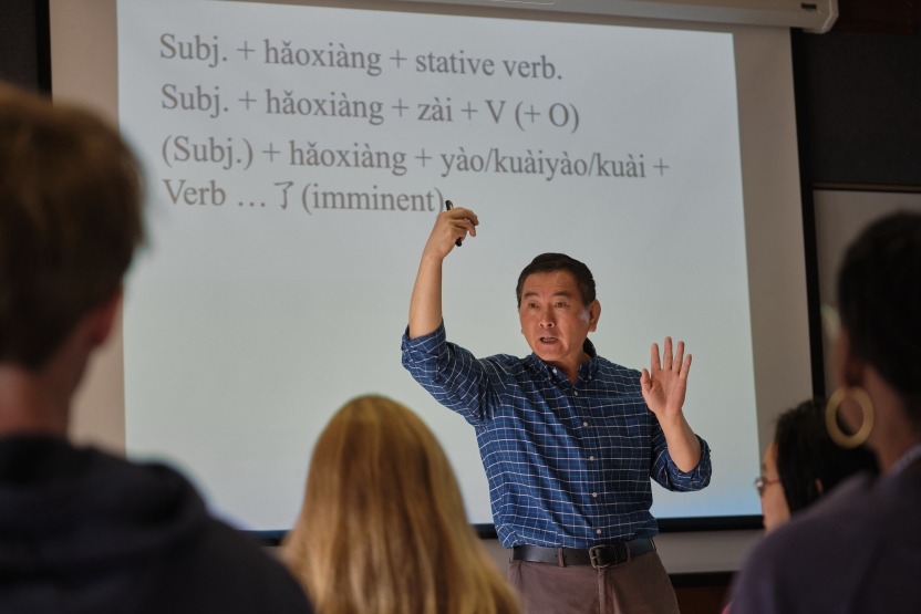 A teacher stands in front of a screen with Chinese characters and a grammar lesson.