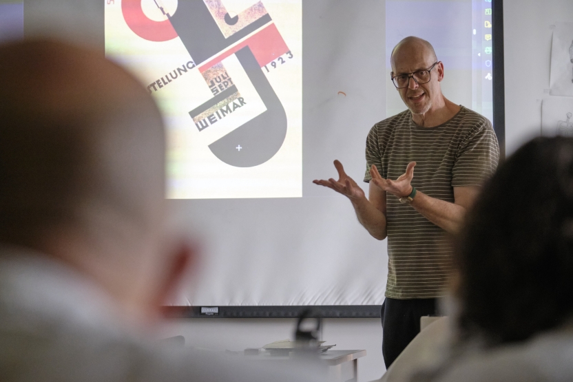 A man stands in front of an illustration on a screen, using his hands to emphasize his point. Students sit in front of him. 