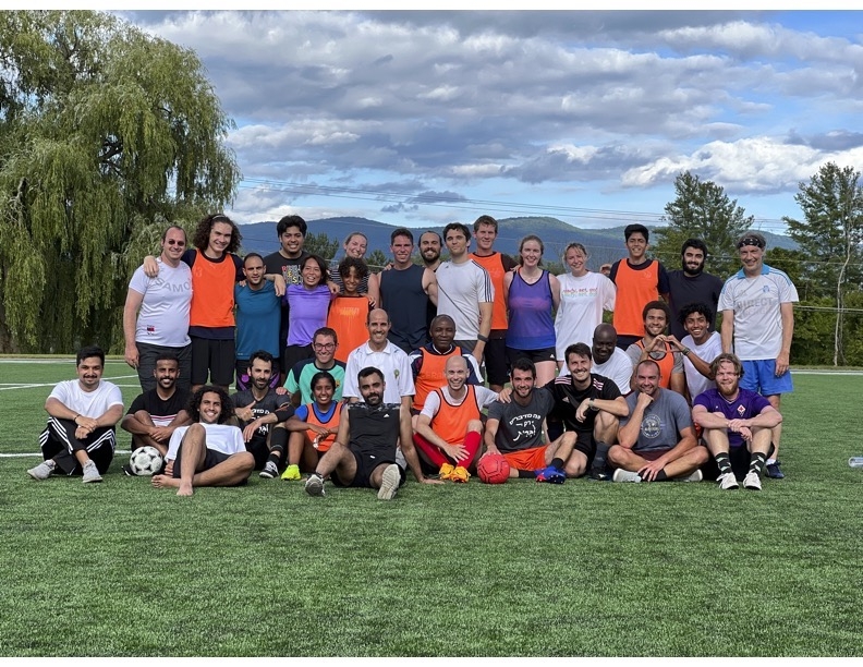 A group of people stands in rows in their exercise clothes after a soccer game, smiling at the camera. 