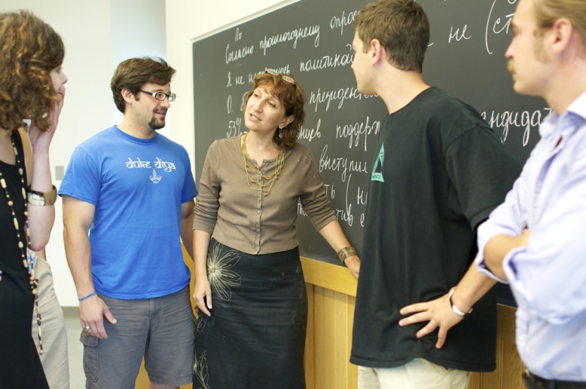 A teacher stands at the blackboard speaking with her students who surround her