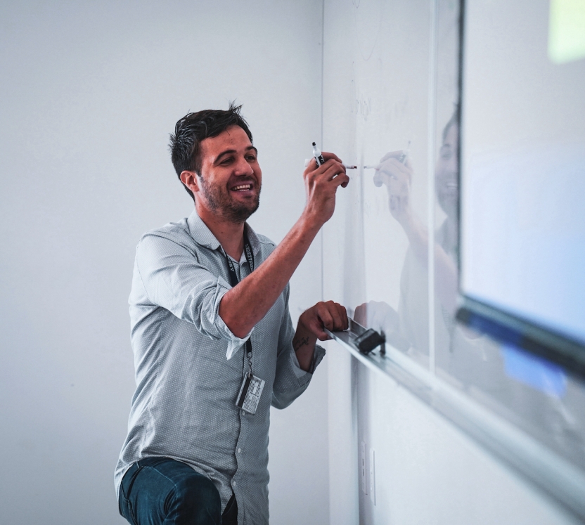 A man writes on a white board, smiling. 