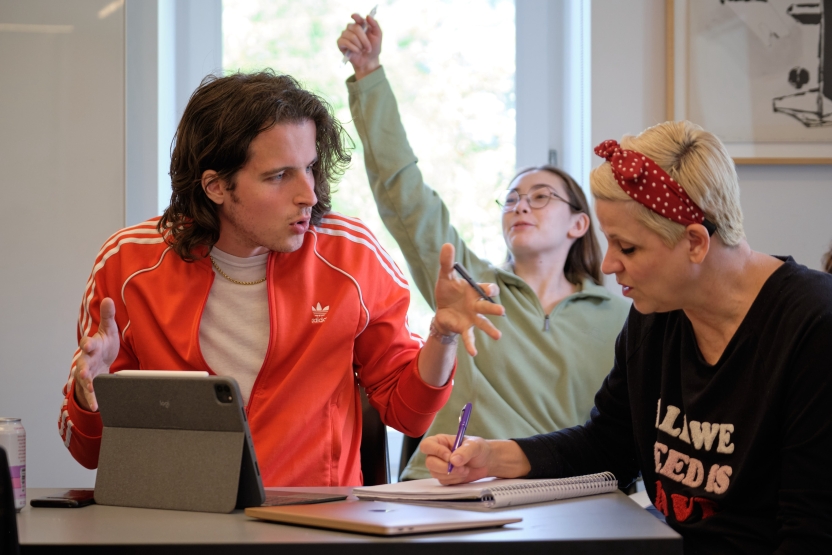 Students discuss a topic at their seats in Italian class. Another student raises her hand behind them.